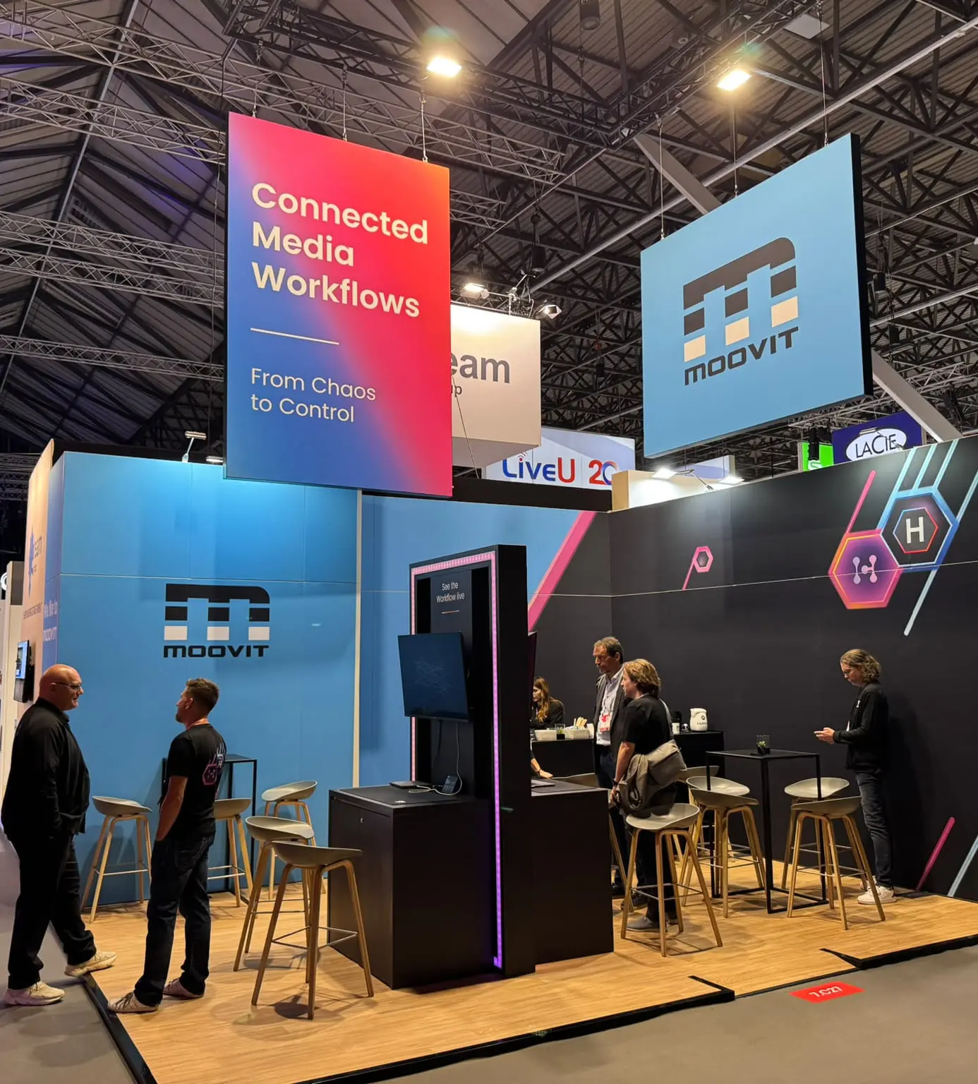 Modulaire beursstand van MoovIT met grote hangende reclamepanelen, zitgedeelte en schermen voor een opvallende presentatie op de beurs.